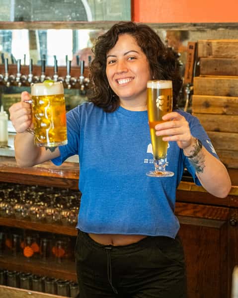 Bartender holding up a large stein and a tall pilsner glass of craft beer inside Künstler Brewing's rustic bar area.