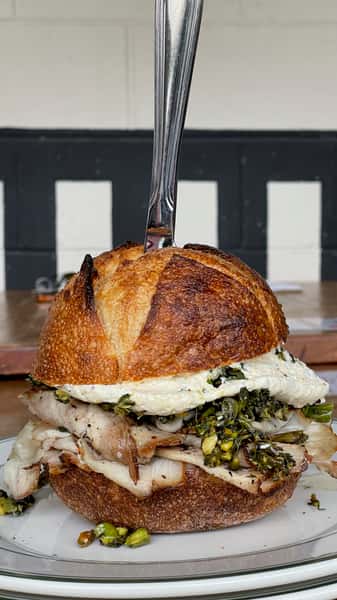 Hearty artisan sandwich stacked with grilled chicken, melted cheese, sautéed greens, and a fried egg, served on rustic bread with a knife through the center.