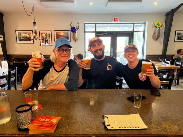 Group of friends smiling and raising craft beers while seated at the bar inside Künstler Brewing, with rustic decor and natural lighting in the background.