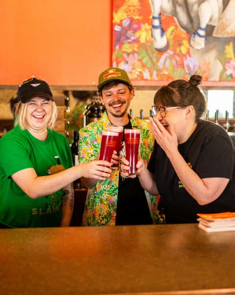 Three friends clinking tall glasses of vibrant red craft beer at Künstler Brewing, laughing together at the colorful bar.
