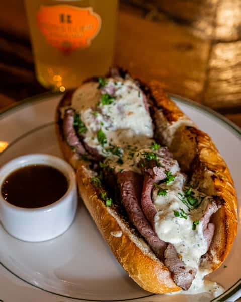 Toasted hoagie roll stuffed with sliced roast beef and topped with creamy horseradish sauce, served with a side of au jus and a Künstler Brewing craft beer.