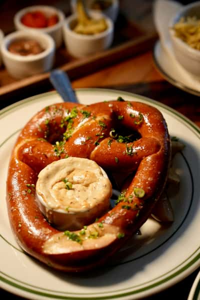 Golden soft pretzel sprinkled with herbs and served with a creamy dipping sauce, plated at Künstler Brewing with German-inspired appetizers in the background.