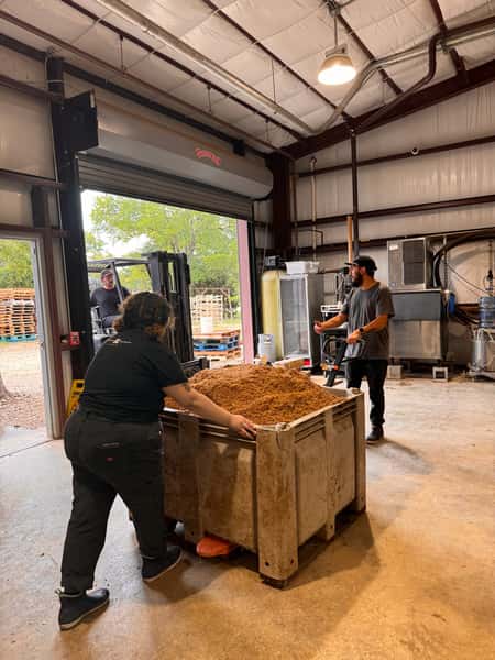 Team at Künstler Brewing moving a large bin of spent grain inside the production facility, with brewing equipment and forklift activity in the background.