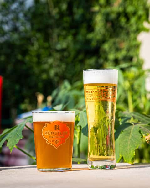Two crisp Künstler Brewing beers—one amber ale and one golden lager—served in branded glasses and set against a lush outdoor backdrop.