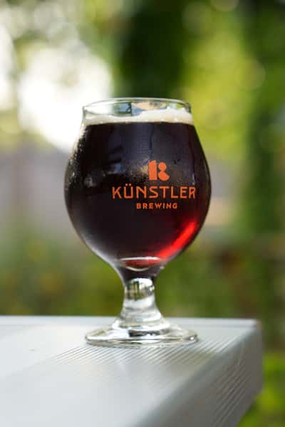 Chilled goblet of dark craft beer from Künstler Brewing, glistening in the sunlight against a blurred green outdoor background.