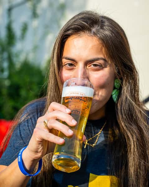Woman sipping from a cold glass of Künstler craft beer while relaxing outdoors on a sunny day, wearing a casual T-shirt and accessories.