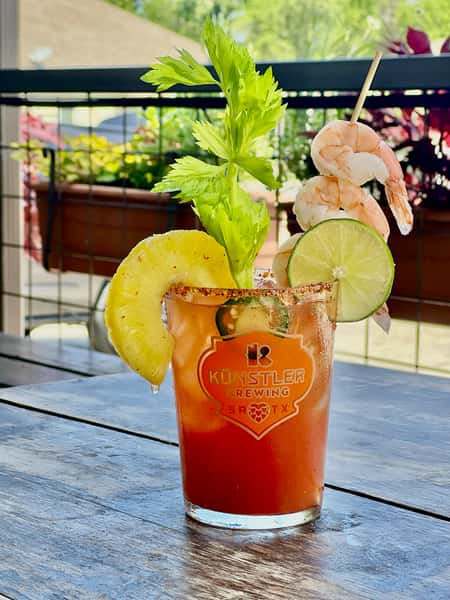 Spicy michelada cocktail from Künstler Brewing in San Antonio, garnished with shrimp, lime, pineapple, celery, and jalapeño on a sunny outdoor patio table.