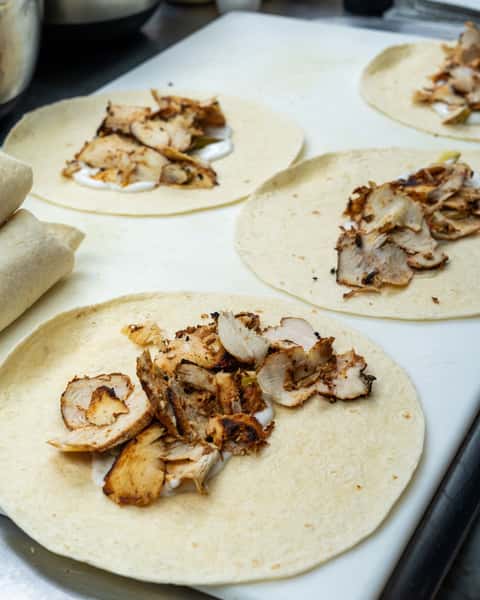 Open tortillas topped with grilled chicken slices and garlic sauce, ready to be wrapped on a kitchen prep counter.