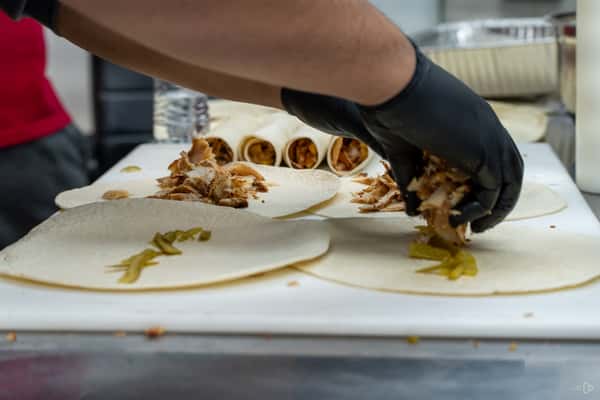 Cook assembling shawarma wraps with grilled meat and pickles on tortillas, wearing black gloves in a kitchen prep station.