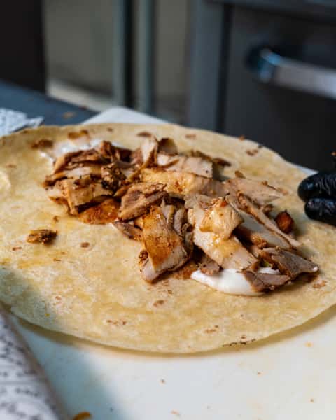 Open tortilla with sliced grilled chicken and white sauce being prepared for a shawarma wrap on a kitchen counter.