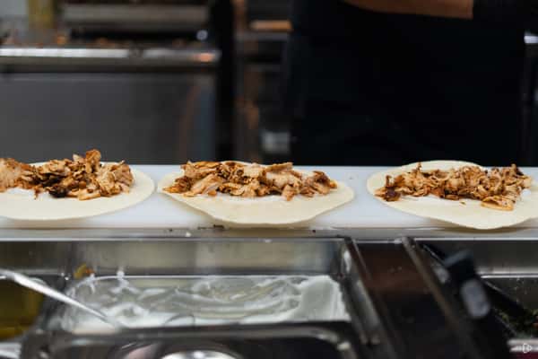 Three open shawarma wraps with grilled chicken lined up on a prep counter beside a toppings station.