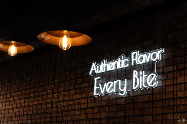 Neon sign on a tiled wall reads "Authentic Flavor Every Bite" under warm hanging lights.