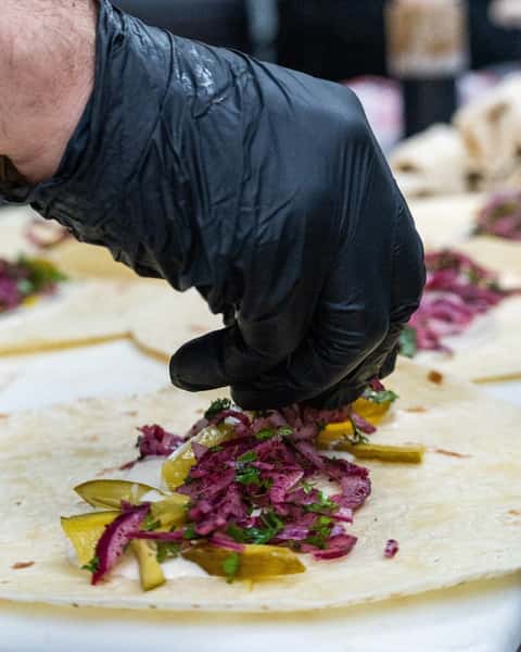 A shawarma wrap being prepared with pickles, purple cabbage, parsley, and white sauce on a flatbread, placed by a gloved hand.