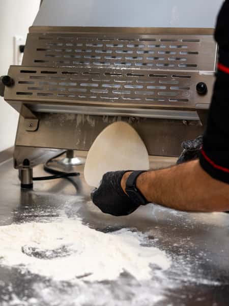 Flattened dough being shaped through a tortilla press on a floured stainless steel surface by a gloved hand.