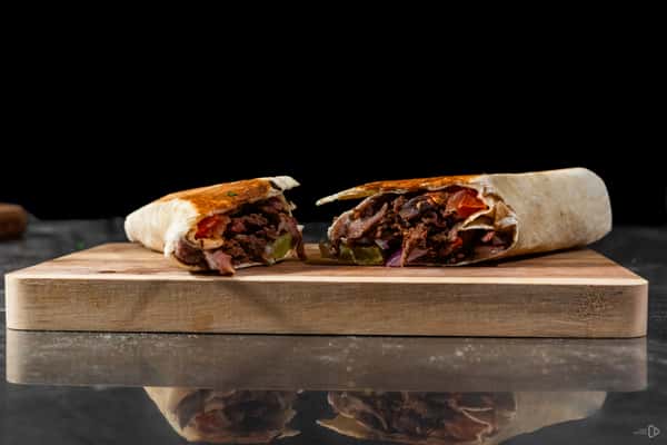 Grilled beef shawarma wrap sliced in half, placed on a wooden board, showcasing juicy meat, onions, pickles, and tomatoes in a toasted tortilla.