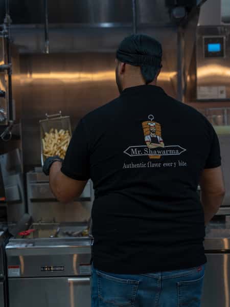 A worker wearing a "Mr. Shawarma" branded shirt is seen from the back, holding a fryer basket of French fries in a commercial kitchen. The shirt reads: "Authentic flavor every bite."