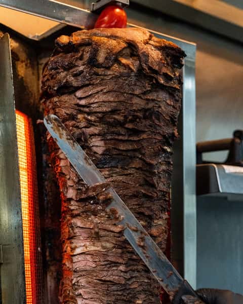 Large rotating spit of beef shawarma being shaved with a long knife beside an open vertical grill.