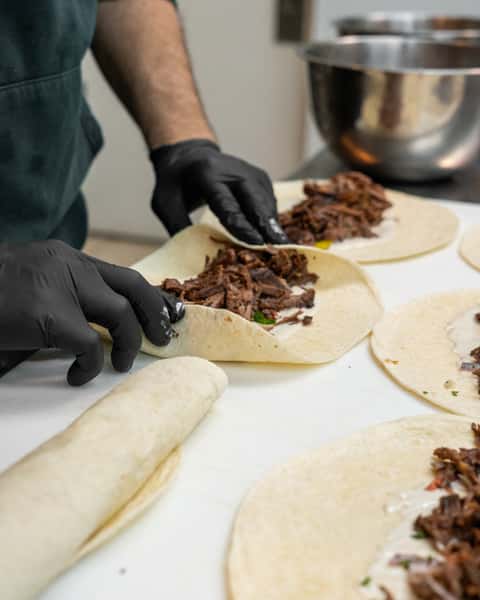 Person wearing black gloves rolling tortillas filled with shredded meat and sauce on a prep counter with metal mixing bowls nearby.