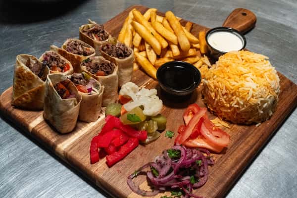 Beef shawarma wrap slices served with fries, rice, pickled vegetables, tomatoes, onions, and dipping sauces on a wooden board.