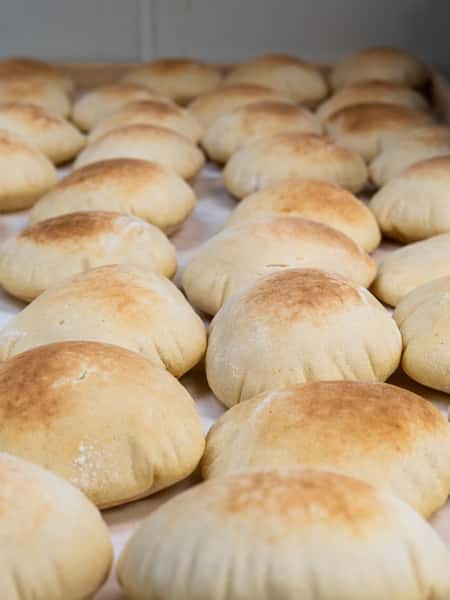 Freshly baked, golden-brown pita breads puffed up with air, cooling on a tray in a bakery or kitchen setting.