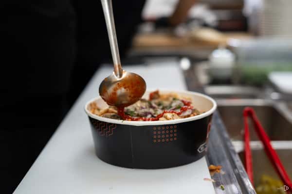 A ladle pours red sauce over a loaded shawarma bowl topped with onions, herbs, and chicken at a fast-casual restaurant.