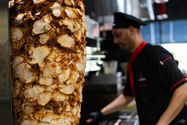 Vertical spit stacked with marinated chicken shawarma meat, with a chef in uniform preparing food in the background.