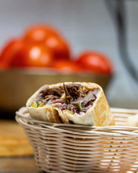 Beef shawarma wrap filled with onions, pickles, and herbs, served in a wicker basket with fresh tomatoes in the background.