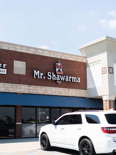 Exterior of Mr. Shawarma restaurant with a branded sign on a brick building and a white SUV parked in front.