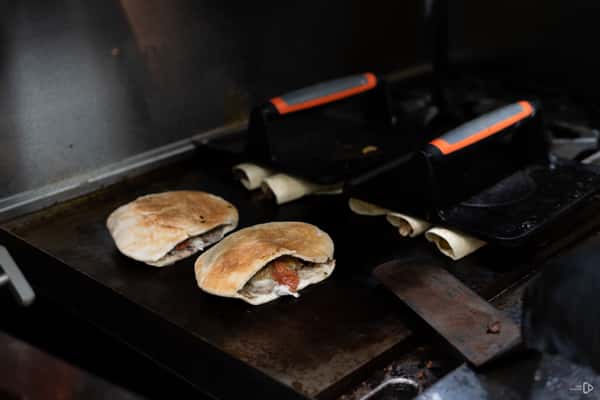 Stuffed pita bread and wraps being grilled on a flat top with heavy presses, partially filled with meat and sauce.