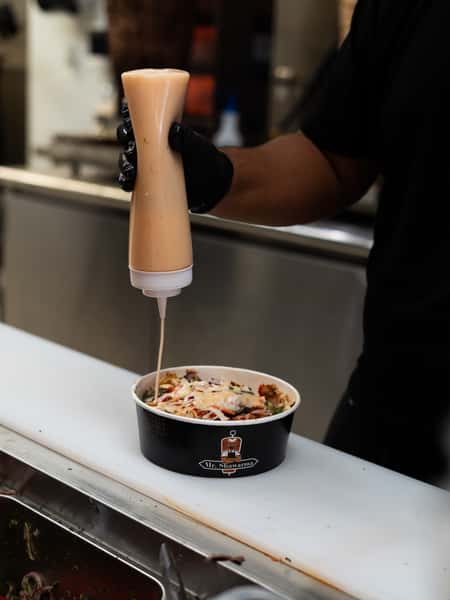 Gloved hand squeezes creamy sauce from a bottle onto a shawarma bowl on a prep counter in a restaurant kitchen.