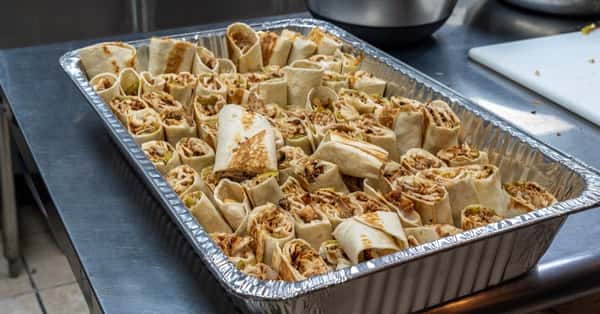 Large aluminum tray filled with neatly arranged grilled chicken shawarma wraps cut into bite-sized pieces, ready for serving.