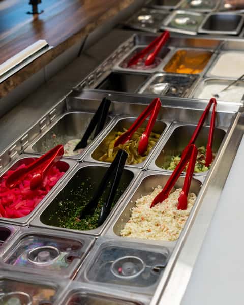 Salad bar with various Middle Eastern toppings like pickled turnips, tabbouleh, jalapeños, coleslaw, and sauces with tongs.
