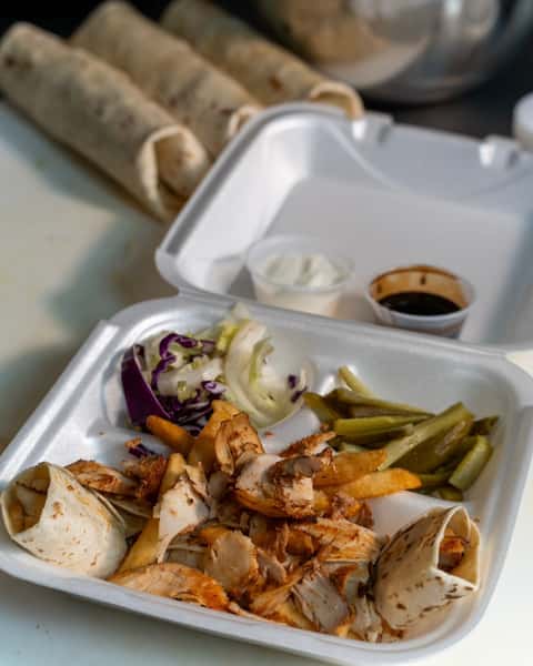 Takeout box with sliced chicken shawarma, fries, pickles, cabbage slaw, wrap pieces, and two sauce containers.