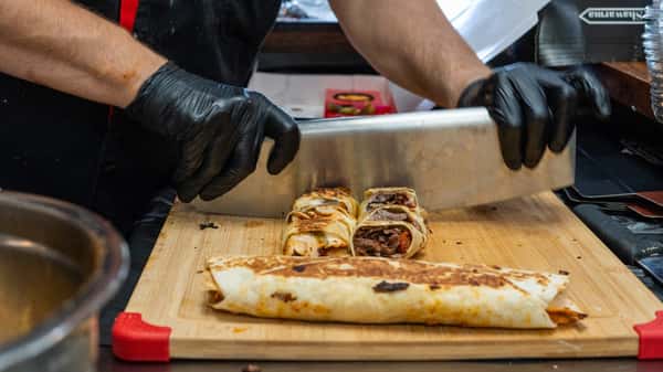 Cook slicing grilled shawarma wraps filled with meat on a wooden board using a large knife, wearing black gloves.