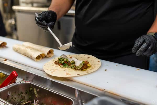 Worker in gloves adds white sauce to a falafel wrap with vegetables on a prep counter near a toppings station.