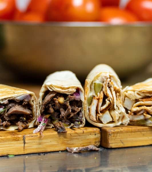 This image shows a close-up of four shawarma wraps cut in half, revealing their fillings. The wraps are placed on a wooden board, with a background featuring a bowl of fresh tomatoes. The fillings appear to be a mix of beef and chicken with visible ingredients like onions, pickles, and possibly garlic sauce or tahini, highlighting the variety and freshness of the shawarma offerings.