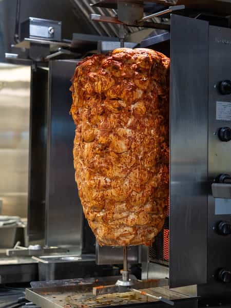 Large stack of marinated chicken shawarma on a vertical rotisserie, cooking evenly in a commercial kitchen setup.