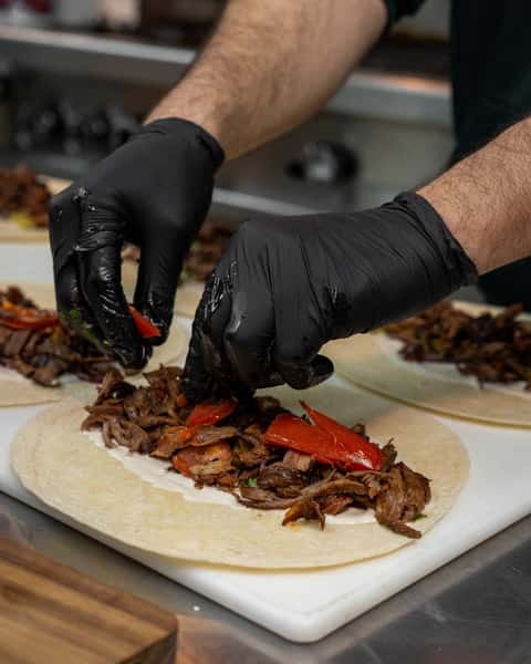 Gloved hands assembling shawarma wraps with sliced beef, tomatoes, and white sauce on flour tortillas in a kitchen setting.