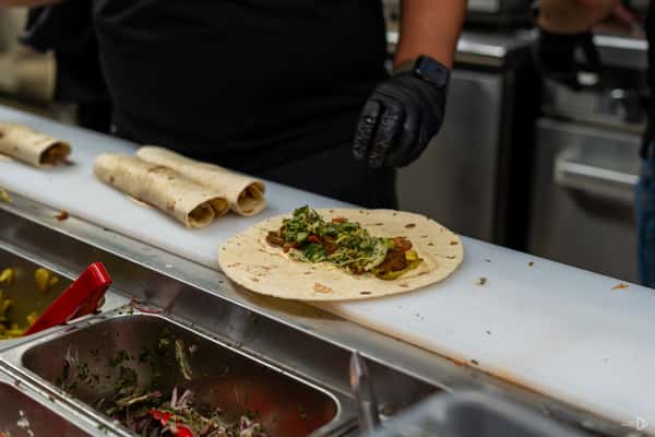 Shawarma wrap being assembled with meat, pickles, and greens on flatbread at a prep counter with rolled wraps nearby.
