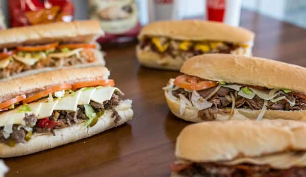 Several steak and cheese sandwiches with various toppings are presented on a wooden surface, suggesting a meal at Steak N' Cheese Please.