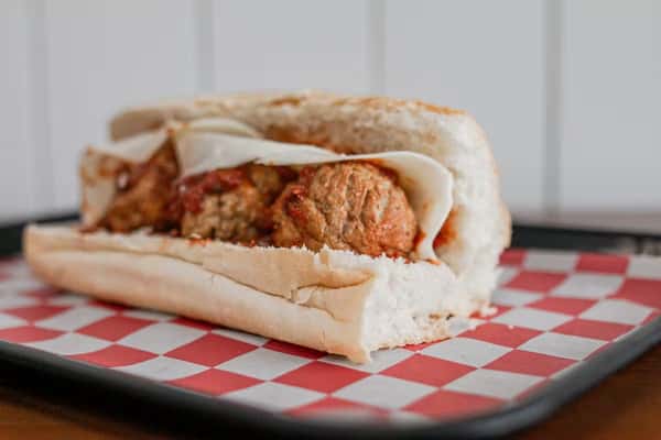 A meatball sub with melted cheese sits on a red and white checkered paper at Steak N' Cheese Please.