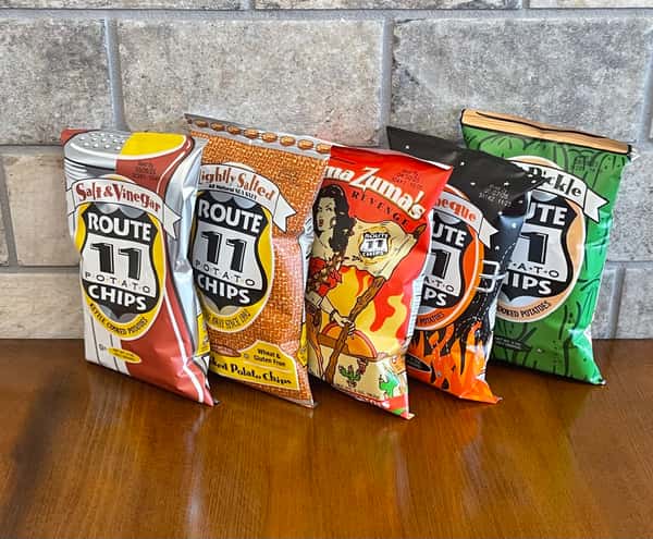 Five bags of Route 11 potato chips in assorted flavors stand on a wooden surface, perhaps ready to be served at Steak N' Cheese Please.