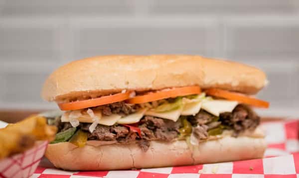 A steak and cheese sub with toppings displayed on a red and white checkered paper at Steak N' Cheese Please.