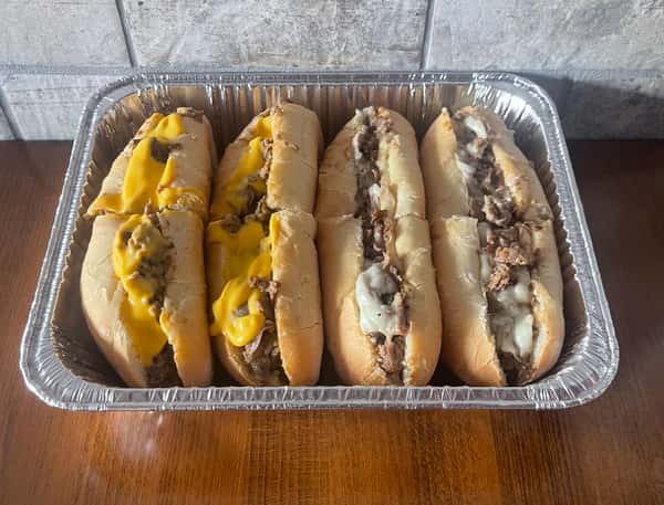 Four steak and cheese sandwiches, some with yellow cheese and others with white cheese, are displayed in a foil pan, ready to be served at Steak N' Cheese Please.