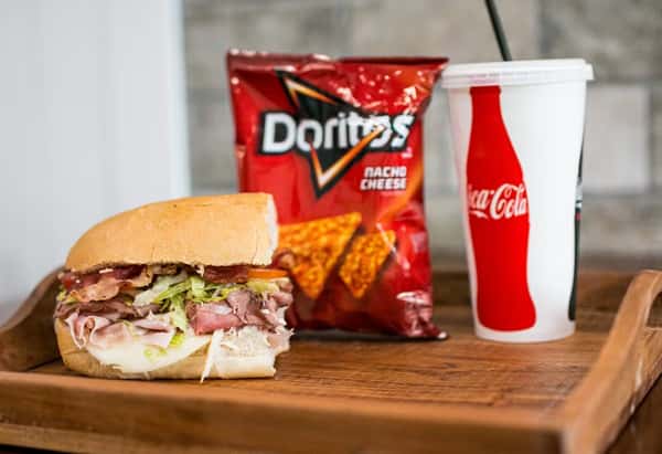 A sandwich, Doritos, and Coca-Cola drink sit on a wooden tray, presenting a meal option from Steak N' Cheese Please.