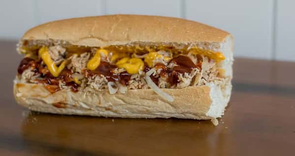 A BBQ chicken sub with cheese and mustard sits on a wooden table from Steak N' Cheese Please.