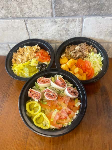 Three salad bowls with varied toppings, including meat, vegetables, and tater tots, are arranged on a table at Steak N' Cheese Please.