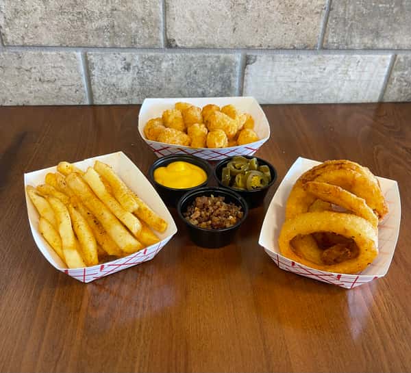 Fries, tater tots, onion rings, cheese sauce, bacon bits, and jalapenos are displayed on a wooden table at Steak N' Cheese Please.