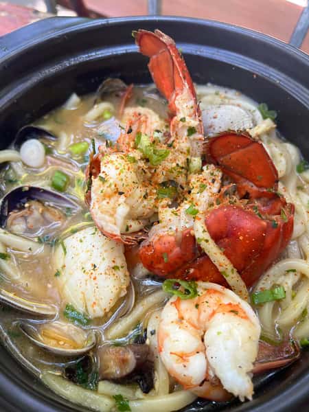 Seafood udon noodle soup with lobster tail, shrimp, clams, and scallops in a savory broth, topped with scallions and seasoning.