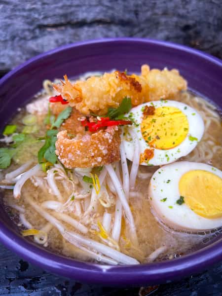 Tempura ramen with crispy shrimp, hard-boiled egg, bean sprouts, herbs, and fried garlic in a rich broth, served in a purple bowl.